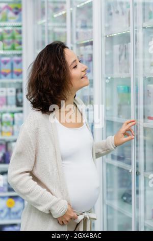 cheerful and pregnant woman with tattoo looking at blurred shelves with medication in drugstore Stock Photo