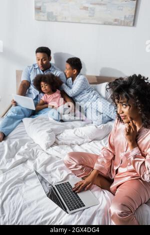 african american woman with laptop looking at blurred husband and kids using digital tablet Stock Photo