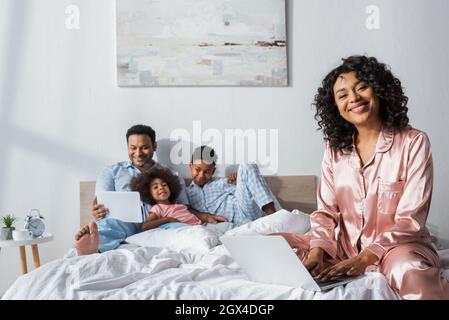 happy african american woman working on laptop near blurred family with digital tablet in bed Stock Photo