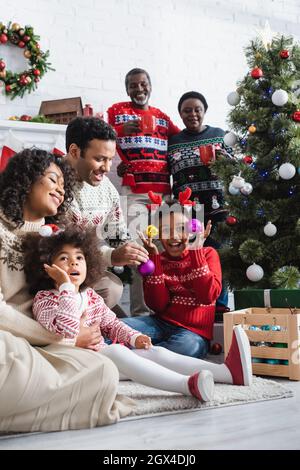 excited boy in reindeer horns headband holding decorative baubles near christmas tree and smiling african american family Stock Photo