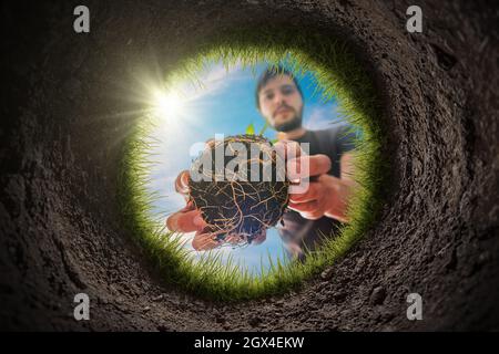 Young man is planting the plant in the garden. View from inside of hole in soil. Stock Photo