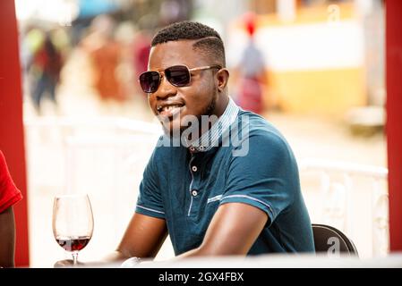 a handsome man sitting in sunglasses in an open air staring at the camera smiling with a glass of red wine. Stock Photo