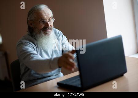 smiling pensive old man with eyeglasses sitting at the desk  and looking at laptop Stock Photo