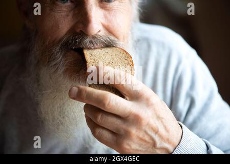 Old sad man with a long gray beard sitting by the table and eating bread Stock Photo