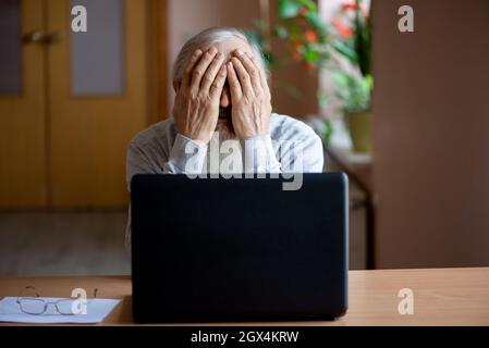 A tired, broken, resigned, preoccupied elderly man sits in front of a laptop, computer and covers his face with his hand. The sad senior checks his ac Stock Photo