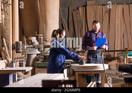 Young female apprentice polishing wood under senior colleague supervision Stock Photo