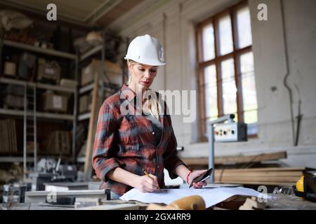 Portrait of female engineer looking at blueprints indoors in carpentry workshop. Small business concept. Stock Photo