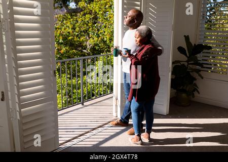 Relaxed african american senior couple drinking coffee standing on balcony in sun Stock Photo