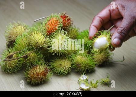 Rambutan fruit pile on wooden background with one peeled fruit held in hand Stock Photo