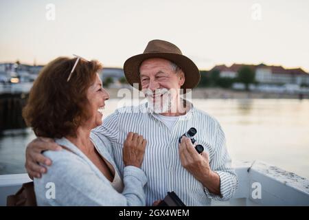 Happy senior couple hugging outdoors on pier by sea, looking at each otherand laughing. Stock Photo