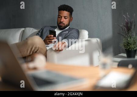 Young african american businessman with smartphone sitting on sofa indoors in office Stock Photo