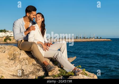 Side view of stylish ethnic couple sitting together on rocks near sea while embracing and enjoying summer Stock Photo