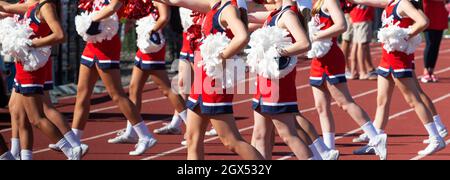 A group of high school cheerleaders cheering on the sidelines oduring a high school football game using red and white pom poms. Stock Photo