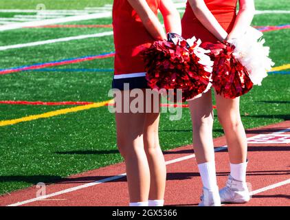 Rear View of two high school cheerleaders holding their pom poms behnd their back during a football game  standing on the track. Stock Photo