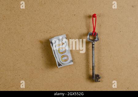 A large metal door key and money against a brown background. The old key hangs vertically on a red rubber band. The dollar bill is attached with a pin Stock Photo