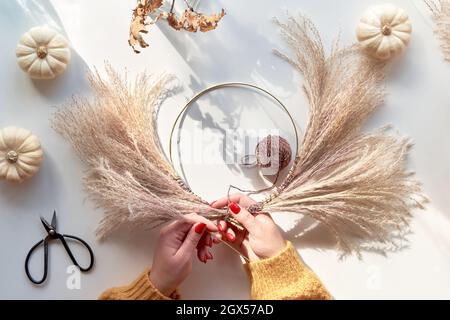 Hands making dried floral wreath from dry pampas grass and Autumn leaves. Hands in sweater with manicured nails tie decorations to metal frame. Top Stock Photo