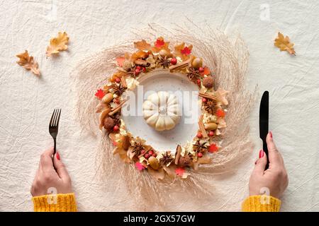 Dry floral wreath on plate with white pumpkin. Hands with fork and knife. Symmetrical central composition. Flat lay, top view on off white tablecloth Stock Photo