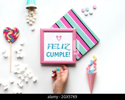 Text Feliz Cumpleanos means Happy Birthday in Spanish. Pink picture frame in hand. Sweets and party decor objects. Candy, snacks, marshmallows Stock Photo