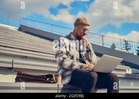 Thoughtful young man using laptop, freelancer working online outdoor, typing on keyboard Stock Photo