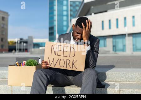 Fired african american businessman with paper poster saying need work and box of personal things, sitting outdoors Stock Photo