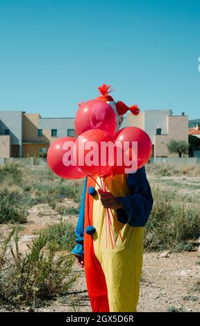 closeup of a creepy clown, wearing a colorful yellow, red and blue costume, is holding a bunch of red balloons in his hand, standing in a vacant lot Stock Photo