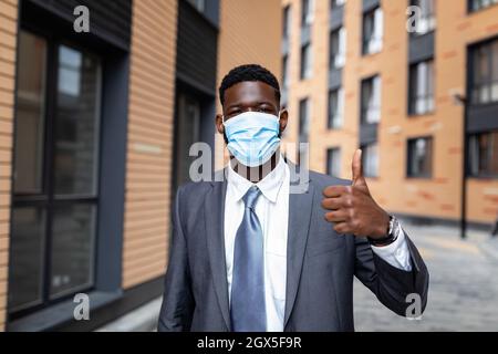 African american businessman in medical face mask and formal outfit, showing thumb up while standing outdoors Stock Photo