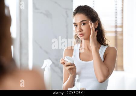 Pretty Female Applying Cream On Face Moisturizing Skin In Bathroom Stock Photo