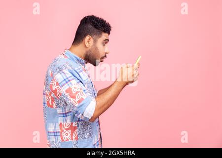 Side view portrait of shocked handsome blogger young adult man with beard wearing in blue shirt using smartphone and watching video with amazed face. Indoor studio shot isolated on pink background. Stock Photo