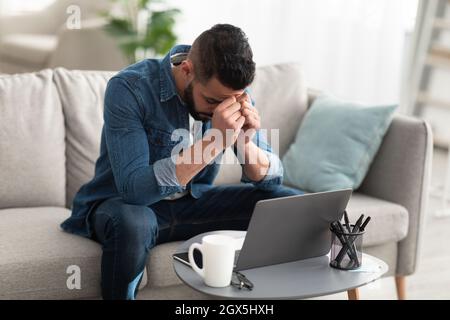 Stressed young Arab man sitting on couch near laptop, having problem with online work or studies at home Stock Photo
