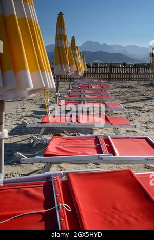 View of empty sand beach-red deck chairs and yellow umbrellas near sea water, Apuan Alps mountains on the horizon and clear sky, Liguria/Toscana Italy Stock Photo