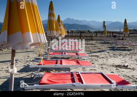View of empty sand beach-red deck chairs and yellow umbrellas near sea water, Apuan Alps mountains on the horizon and clear sky, Liguria/Toscana Italy Stock Photo