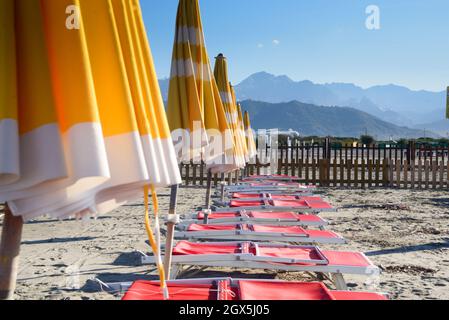 View of empty sand beach-red deck chairs and yellow umbrellas near sea water, Apuan Alps mountains on the horizon and clear sky, Liguria/Toscana Italy Stock Photo