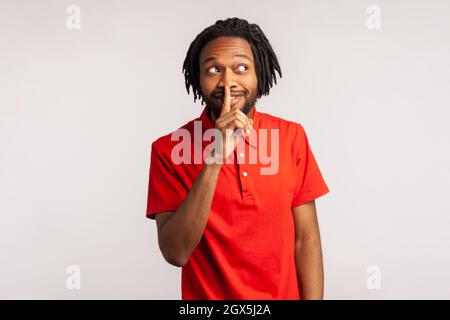 Positive bearded man with dreadlocks wearing red casual style T-shirt, showing hush with finger on his lips gesture, shushing, asking to keep silence. Indoor studio shot isolated on gray background. Stock Photo