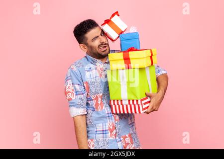 Portrait of funny bearded man carrying lot of heavy present boxes falling on him, looking with frowning face, trying to hold many birthday gifts. Indoor studio shot isolated on pink background. Stock Photo