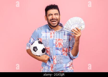 Extremely excited happy man with beard in blue casual shirt holding soccer ball and dollar bills, screaming, betting and winning, keeps eyes closed. Indoor studio shot isolated on pink background. Stock Photo