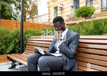African american businessman in suit reading newspaper while sitting on bench near office center, free space Stock Photo