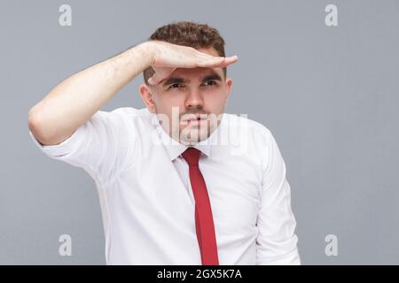 Portrait of young handsome attentive man in white shirt and tie standing with hand on forehead and looking far with attentive face. indoor isolated on gray background. Stock Photo