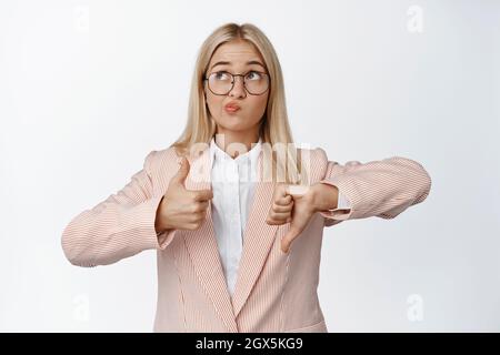 Saleswoman making decision, showing thumbs up and down, thinking how to avaluate, standing over white background Stock Photo