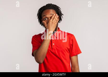 Portrait of young bearded man with dreadlocks wearing red casual style T-shirt, spying through hole in fingers closing eyes with palm, peeking. Indoor studio shot isolated on gray background Stock Photo