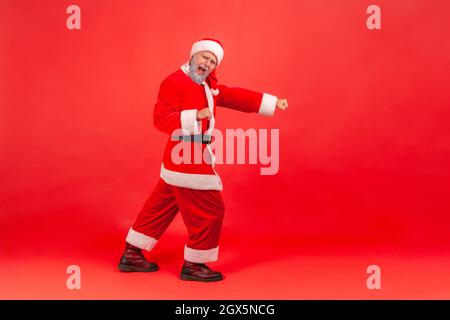 Full length portrait of positive elderly man with gray beard in santa claus costume moving happily, celebrating great creative idea, excited expression. Indoor studio shot isolated on red background. Stock Photo