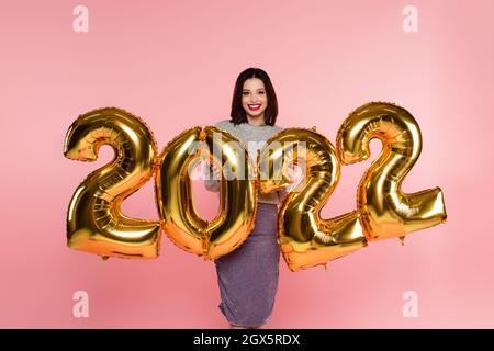 Happy stylish woman standing near balloons in shape of 2022 numbers isolated on pink