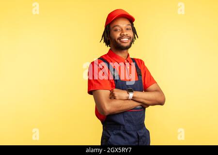 Satisfied handyman wearing overalls standing with crossed arms, looking at camera, profession of service industry, courier delivery. Indoor studio shot isolated on yellow background. Stock Photo