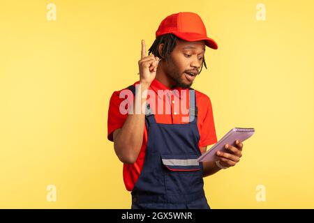 Excited worker wearing blue overalls and red cap holding paper notebook, writing down orders, having new idea of service, pointing finger up. Indoor studio shot isolated on yellow background. Stock Photo