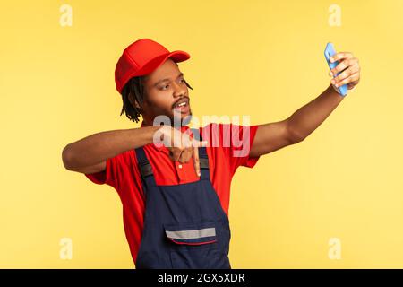 Handyman wearing blue uniform having video call with client, pointing down, asking to subscribe on social network of his service industry. Indoor studio shot isolated on yellow background. Stock Photo