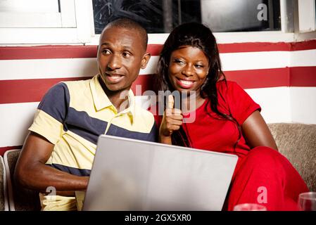 beautiful young african couple sitting indoors and working using a laptop computer while being happy, beautiful woman making a sign of ok with his thu Stock Photo