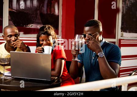 young people sitting at table in a restaurant outside talking while working with a laptop computer and drinking a glass of red wine each Stock Photo