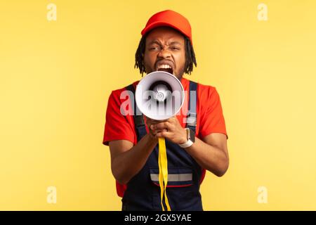 Portrait of angry bearded worker wearing red T-shirt and blue overalls holding megaphone and screaming with aggressive expression, protesting. Indoor studio shot isolated on yellow background. Stock Photo