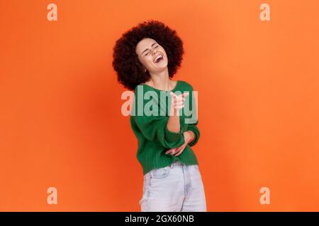 Woman with Afro hairstyle wearing green sweater holding hand on stomach and laughing out loud, pointing finger to camera, indicating ridiculous idiot. Indoor studio shot isolated on orange background. Stock Photo