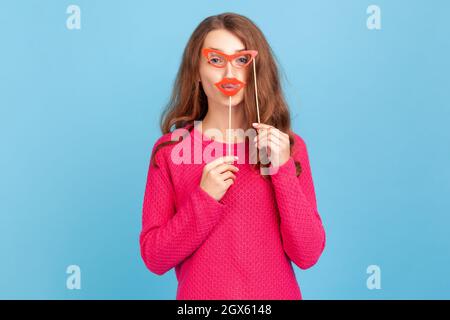 Portrait of positive woman in pink pullover, holding paper glasses and lips, looking at camera, having festive mood, wearing party props. Indoor studio shot isolated on blue background. Stock Photo