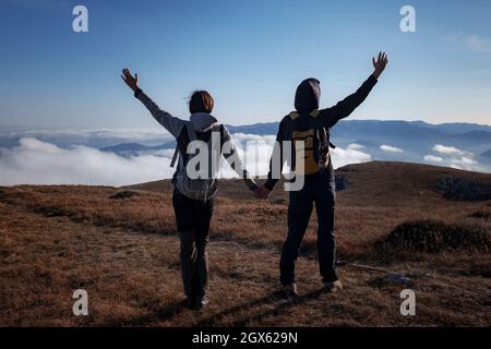 Hiking adventure healthy outdoors people standing talking. Couple enjoying view above clouds on trek. young woman and man in nature wearing hiking backpacks and sticks. Stock Photo